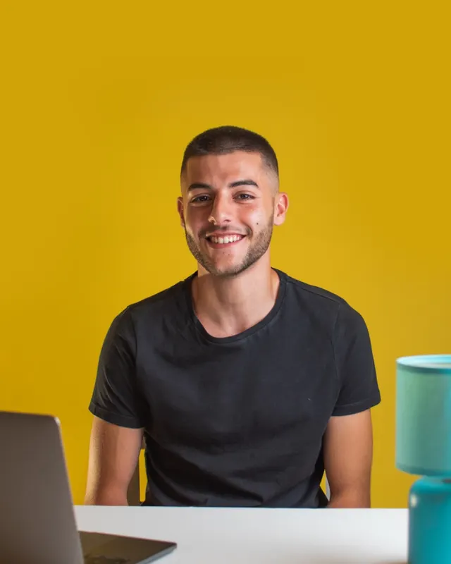 Stephen smiling at a desk with a laptop against a yellow wall, June 2021