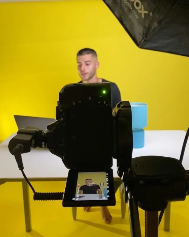 A camera on a tripod filming Stephen at a desk against a yellow wall, June 2021